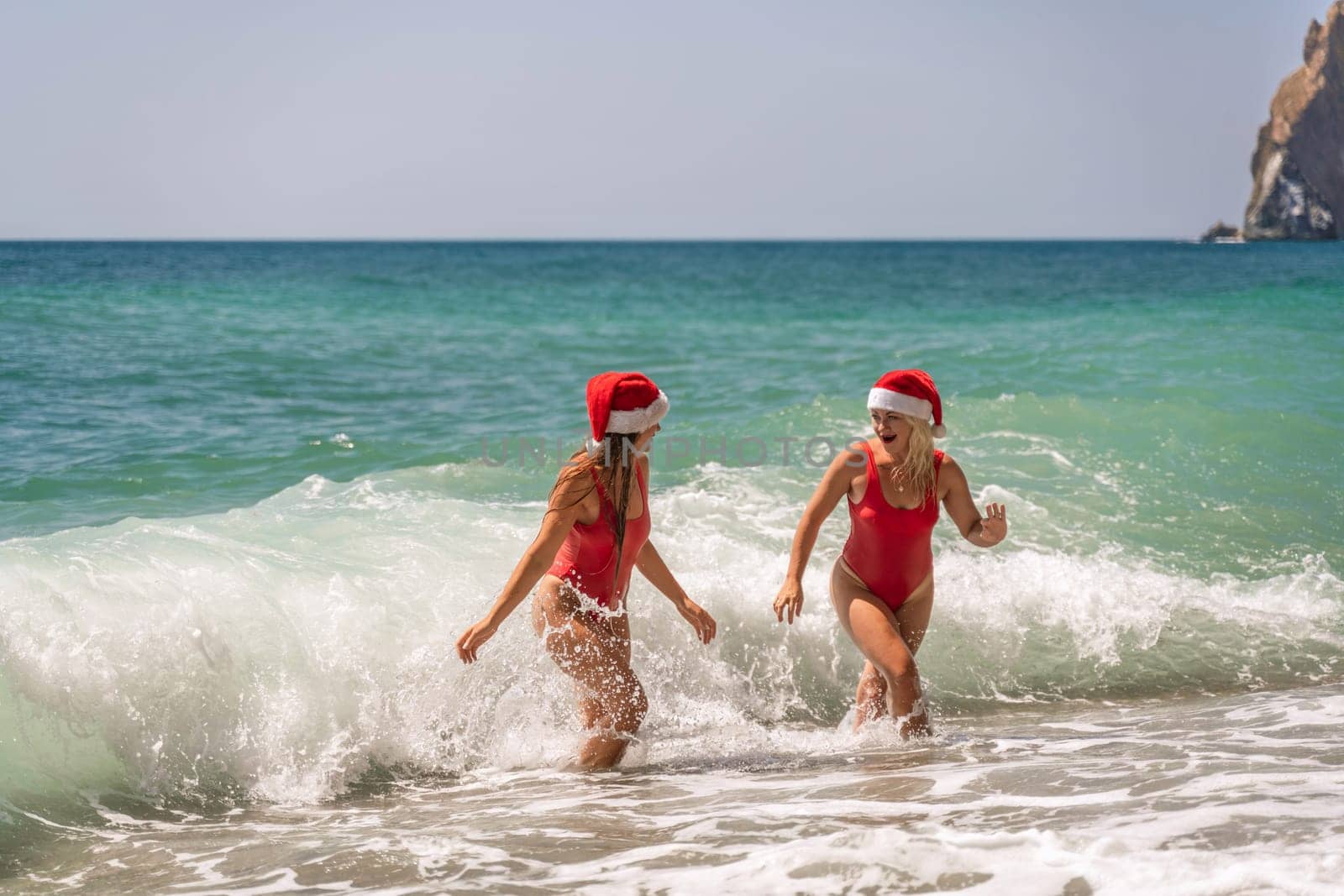 Women Santa hats ocean play. Seaside, beach daytime, enjoying beach fun. Two women in red swimsuits and Santa hats are enjoying themselves in the ocean waves