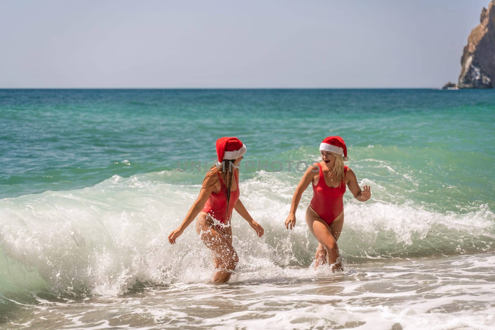 Women Santa hats ocean play. Seaside, beach daytime, enjoying beach fun. Two women in red swimsuits and Santa hats are enjoying themselves in the ocean waves. by Matiunina