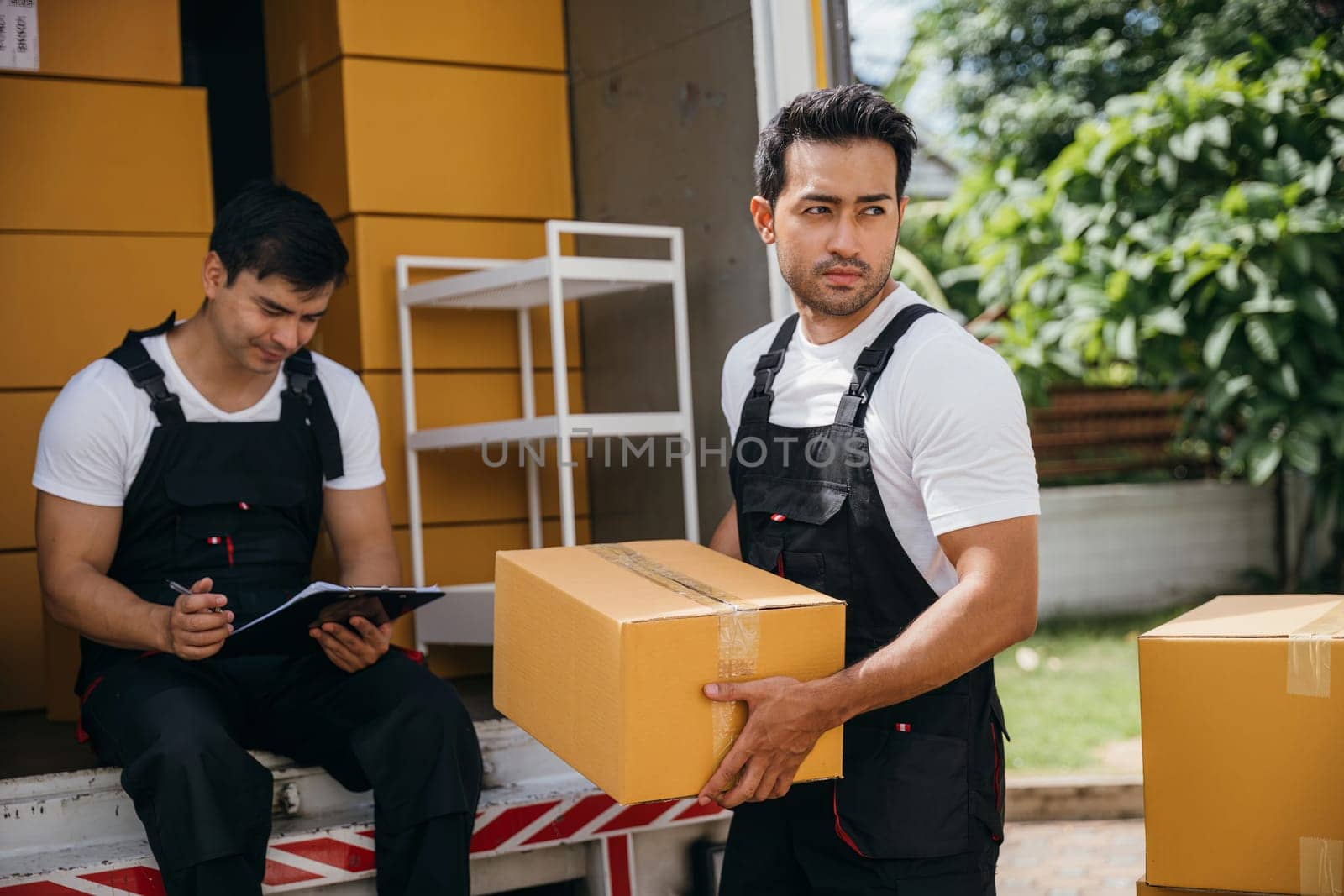 Mover workers in uniform unload boxes checking shipment with a clipboard at the truck. Expert movers ensure efficient moving and professional service. Moving day concept by Sorapop