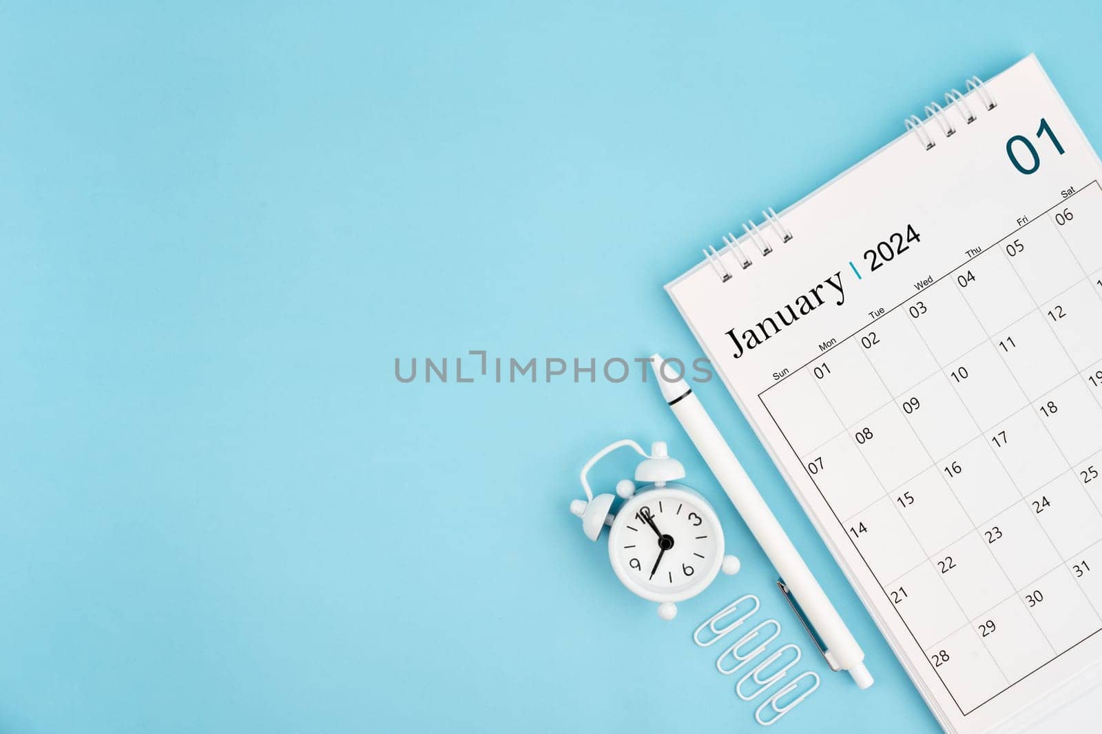 January 2024, Monthly desk calendar for 2024 year and alarm clock with paper clips on blue color background. by Gamjai