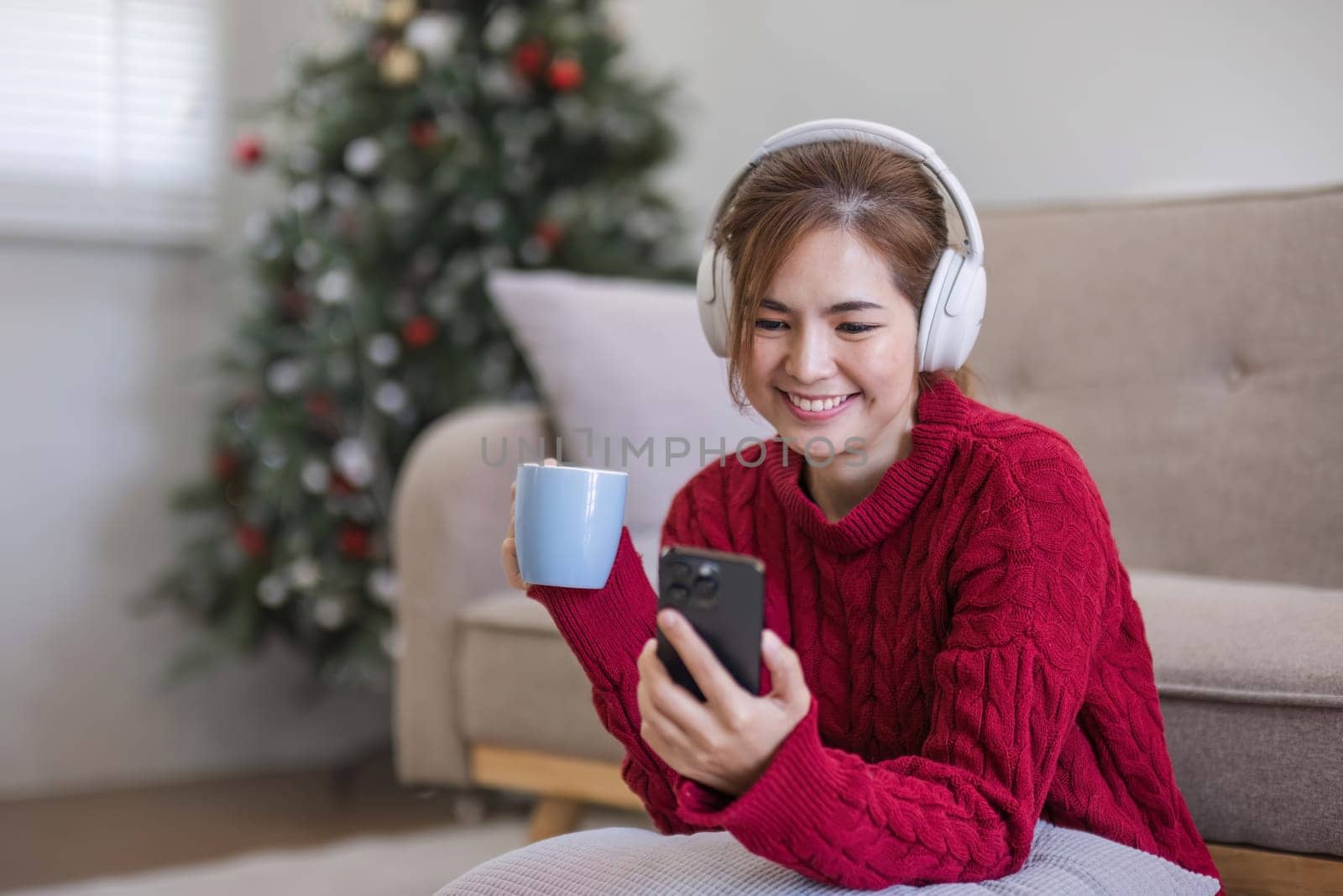 Asian woman sitting on sofa in living room using smartphone and listening to music on headphones..