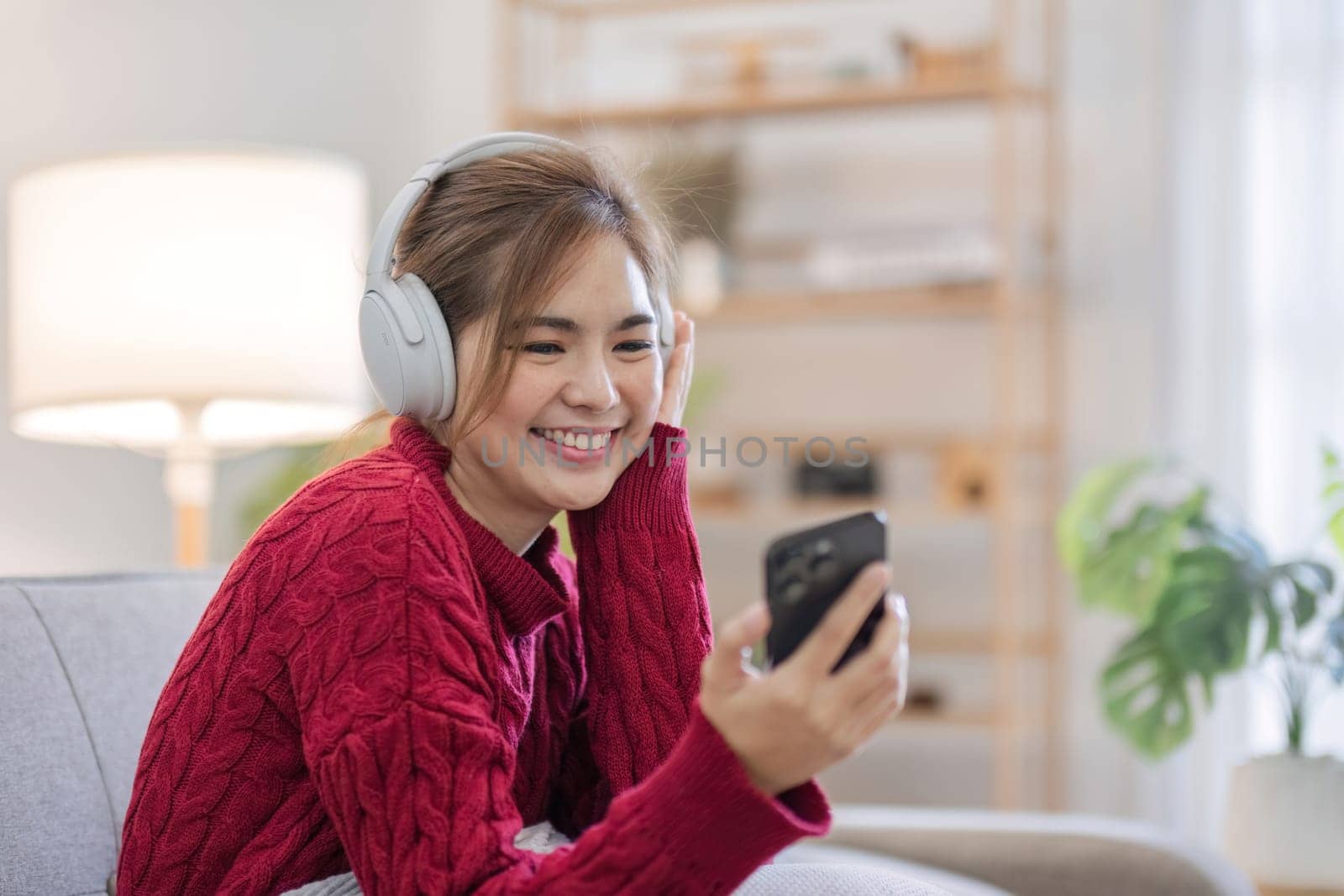 Asian woman sitting on sofa in living room using smartphone and listening to music on headphones. by wichayada