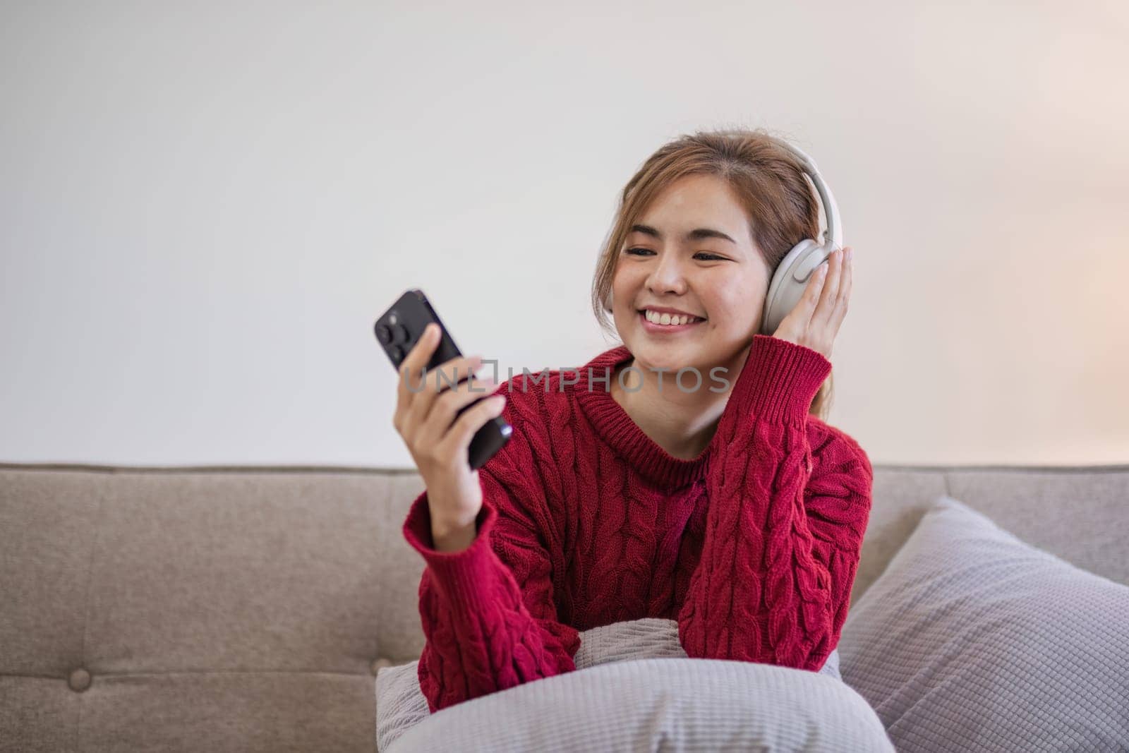 Asian woman sitting on sofa in living room using smartphone and listening to music on headphones. by wichayada