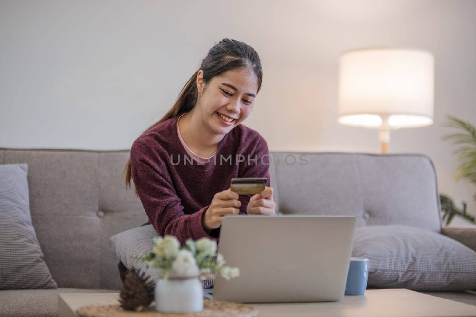 Young Asian woman in a good mood sits on the sofa in the living room and uses a laptop and a credit card. Check out the new website for online shopping. by wichayada