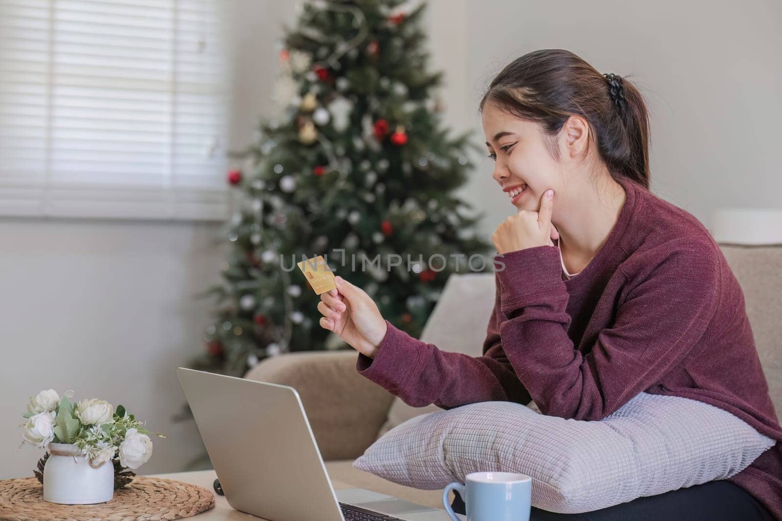 Young Asian woman in a good mood sits on the sofa in the living room and uses a laptop and a credit card. Check out the new website for online shopping. by wichayada