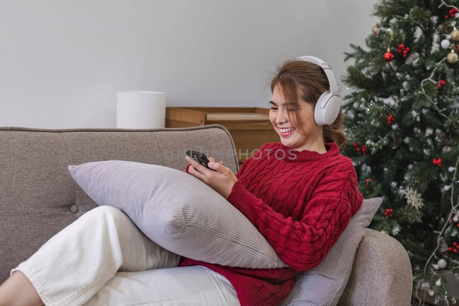 Asian woman sitting on sofa in living room using smartphone and listening to music on headphones. by wichayada