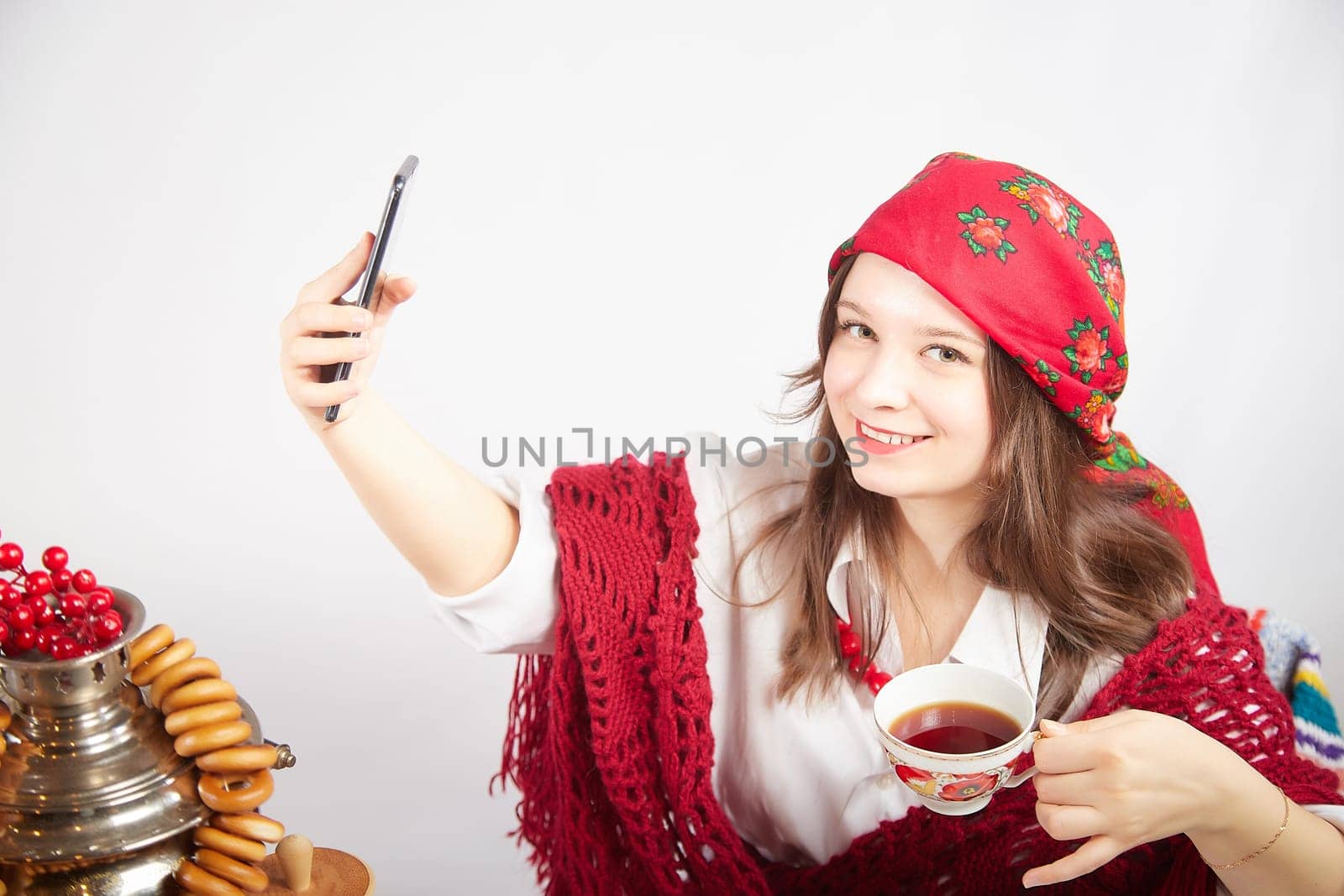 A fashionable modern girl in stylized folk clothes at a table with a samovar, bagels and tea takes a selfie on the Orthodox holiday of Maslenitsa and Easter. Funny photo shoot for a young woman by keleny