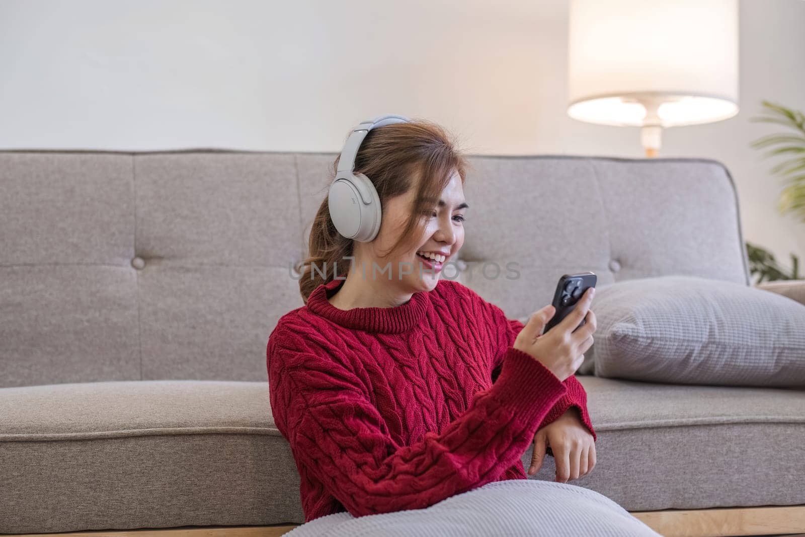 Asian woman sitting on sofa in living room using smartphone and listening to music on headphones. by wichayada