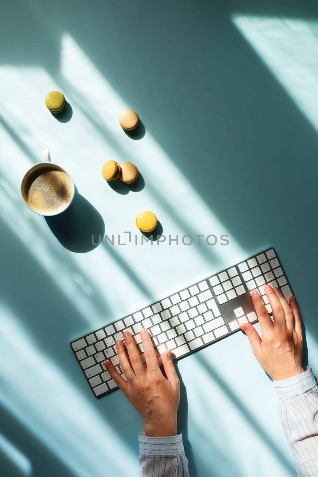 Workplace at home by the window. Flat lat with cup of coffee and macaroons in sunlight and women hands typing on the keyboard.