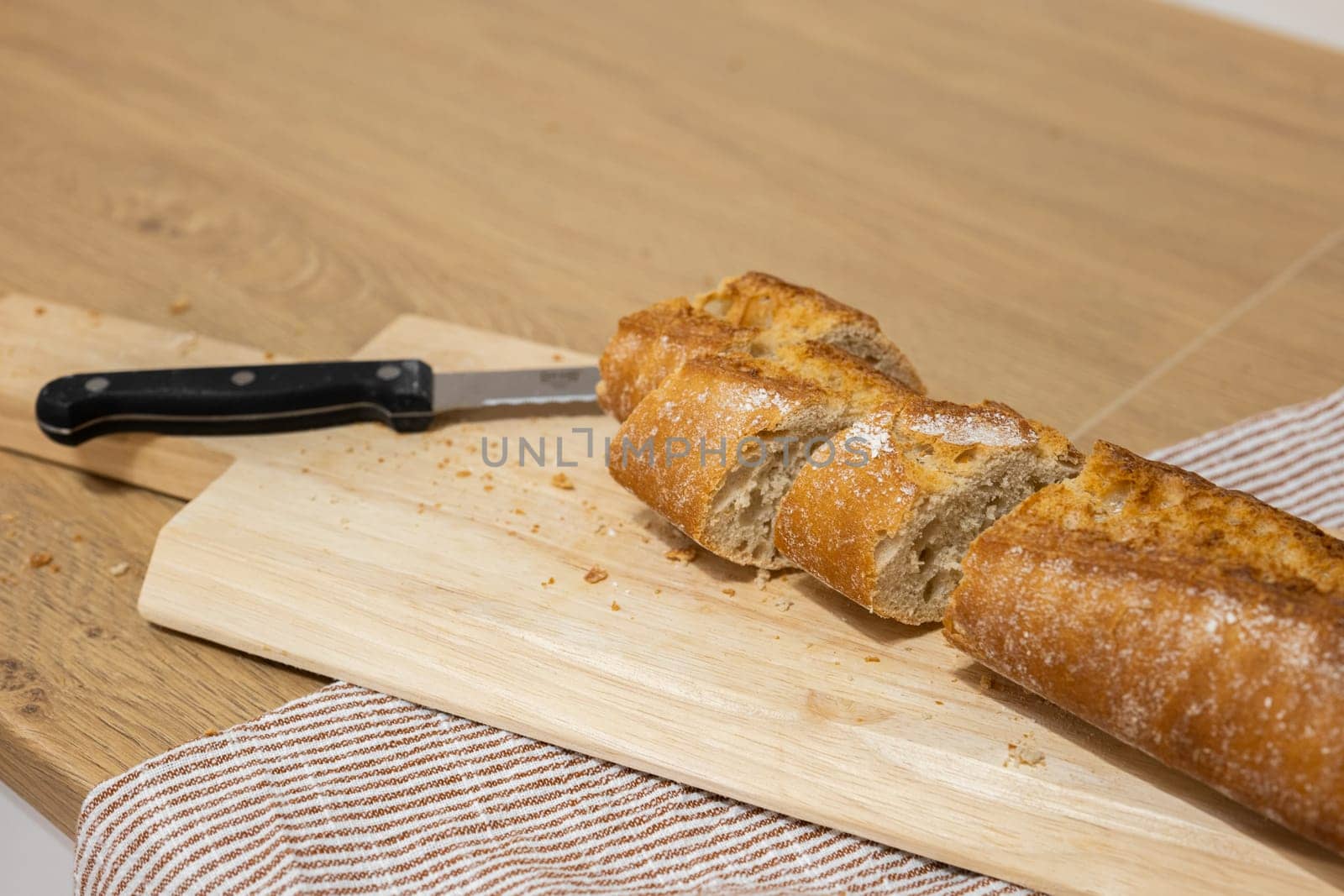 Sliced pieces of French bread with knife on cutting board put on kitchen towel. Process of making sandwiches in modern kitchen. Kitchen ware