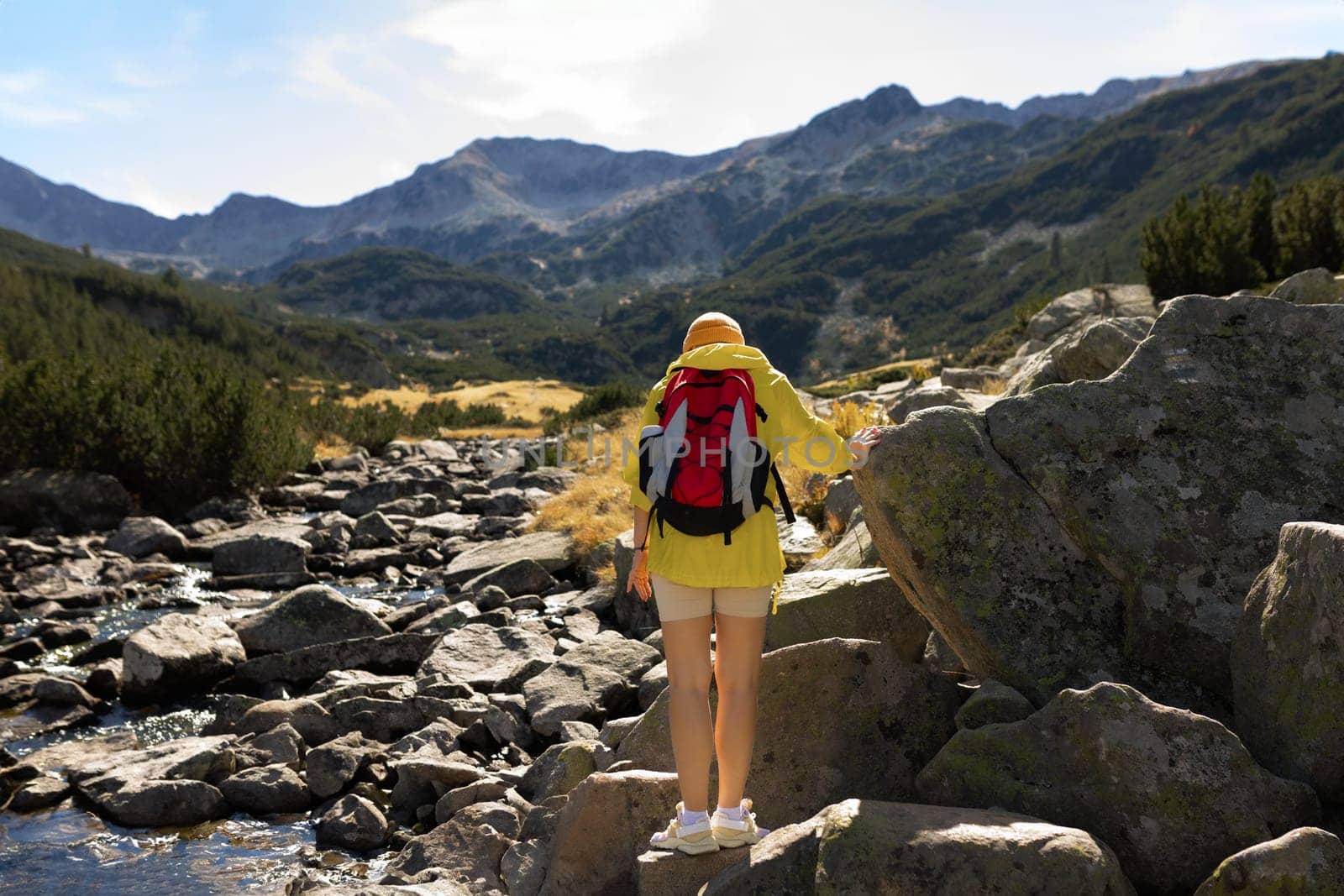 A girl on top of a mountain with green grass looks at a beautiful mountain valley. Landscape with a sporty young woman in a yellow raincoat. Travel and tourism. Hiking.