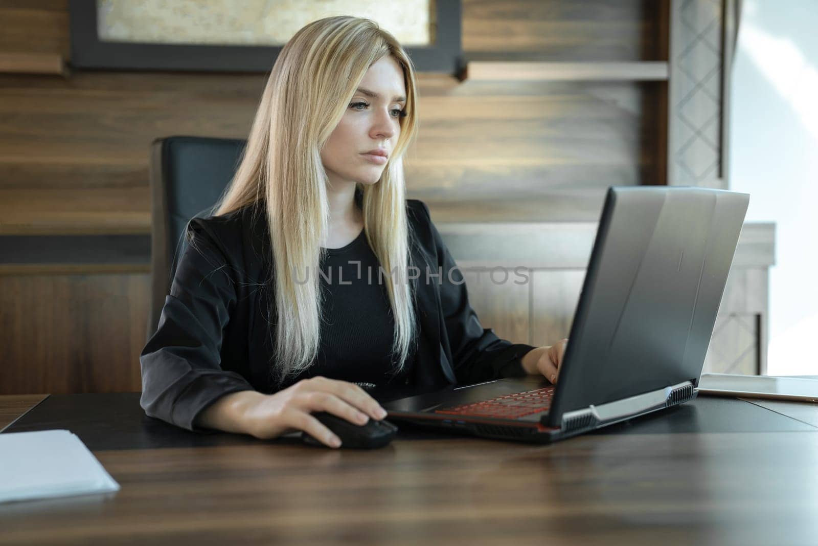 Blonde girl in business clothes sits in front of laptop in office and works intently, concept of online education and work.