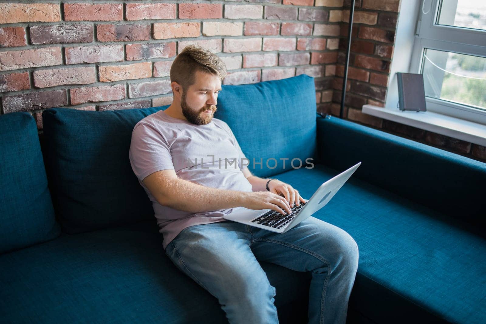 Millennial man sitting on sofa at home working on laptop online and using internet. Smiling and happy mood, freelancer and free leisure time, relaxed and modern job lifestyle concept. Copy space and empty place for text by Satura86