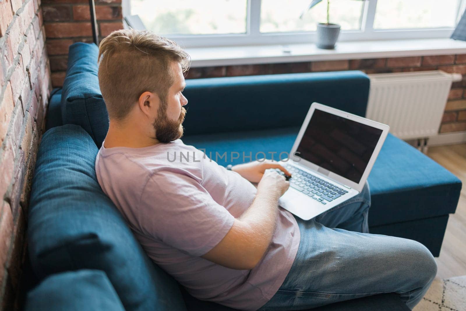 Millennial man sitting on sofa at home working on laptop online and using internet. Smiling and happy mood, freelancer and free leisure time, relaxed and modern job lifestyle concept. Copy space and empty place for text by Satura86