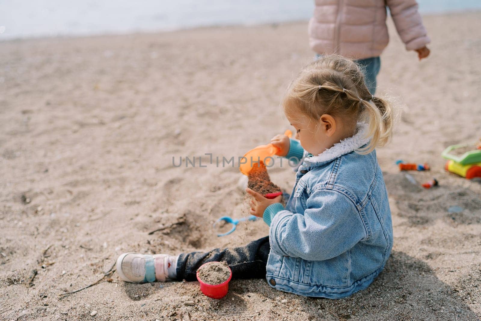 Little girl sits on the beach and pours sand with a shovel into a plastic mold. High quality photo