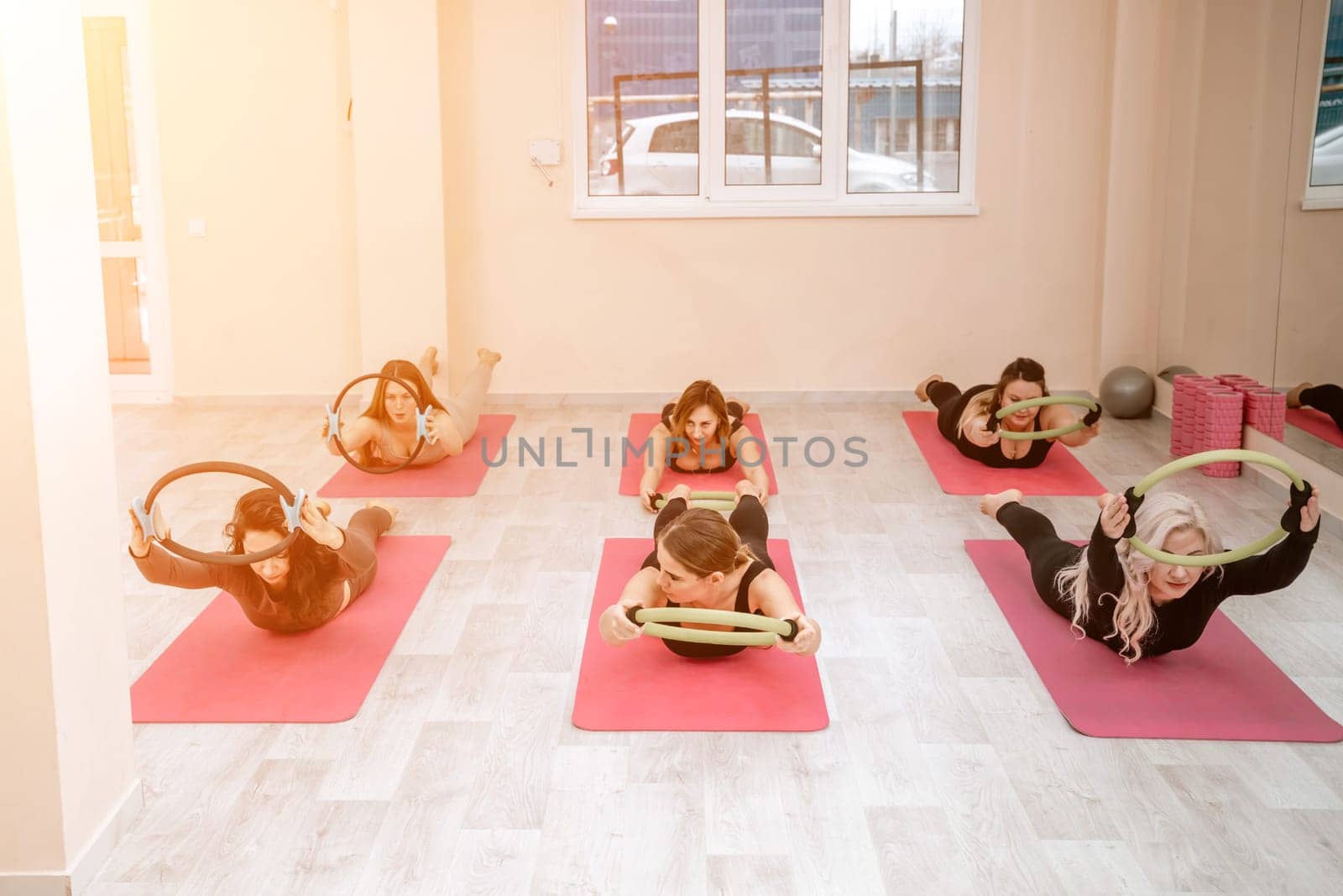 A group of six athletic women doing pilates or yoga on pink mats in front of a window in a beige loft studio interior. Teamwork, good mood and healthy lifestyle concept. by Matiunina