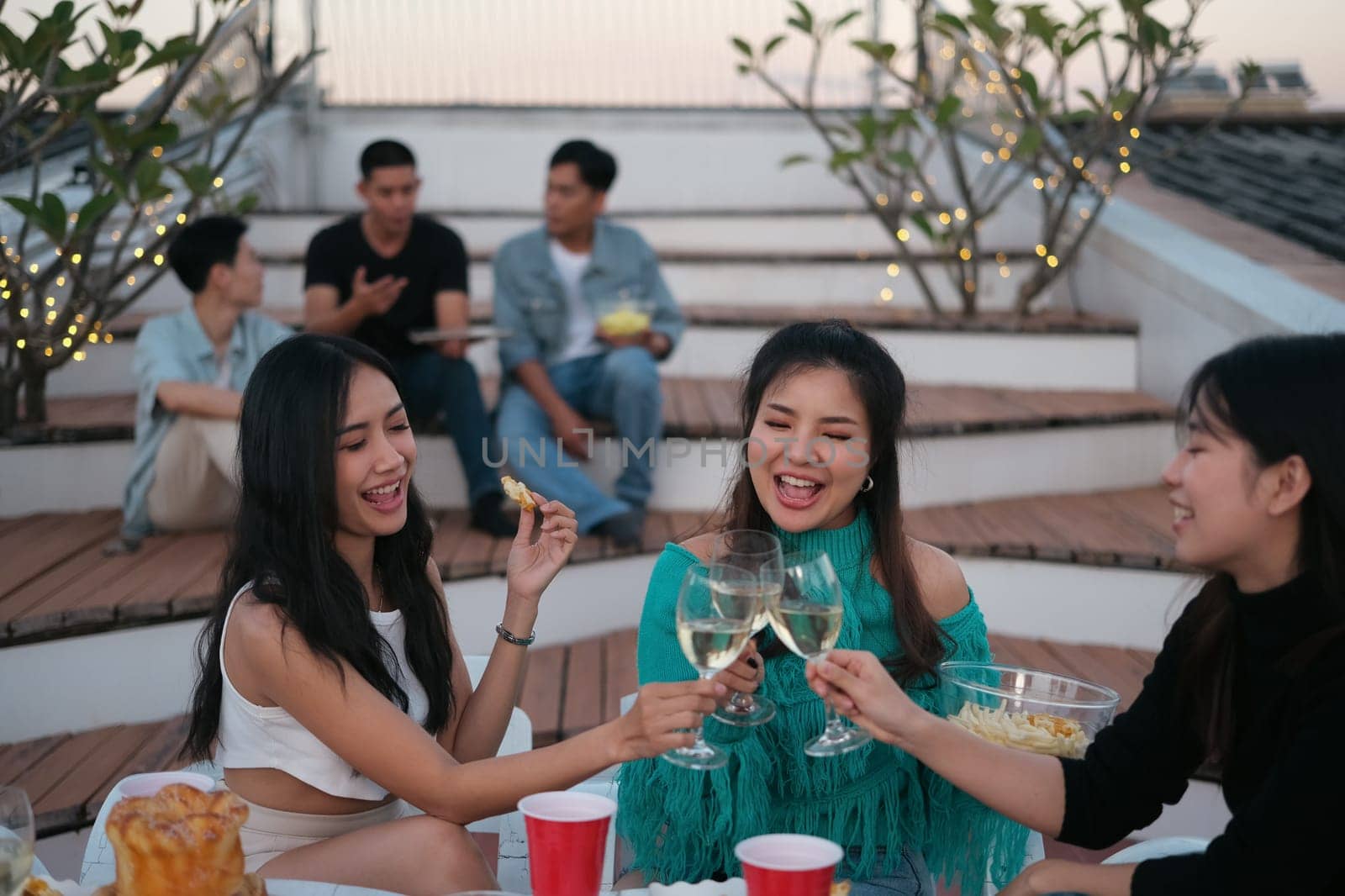 Cheerful male and female friends enjoying happy hour at terrace party and toasting drinks