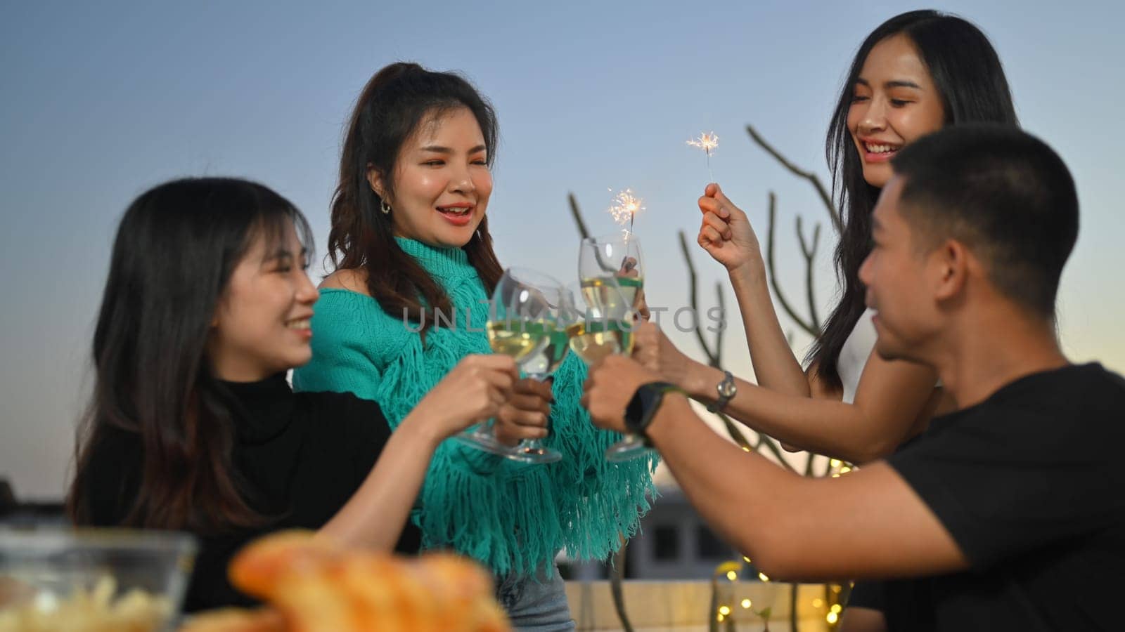 Happy female and male friends igniting sparkler and toasting drinks on building terrace at night