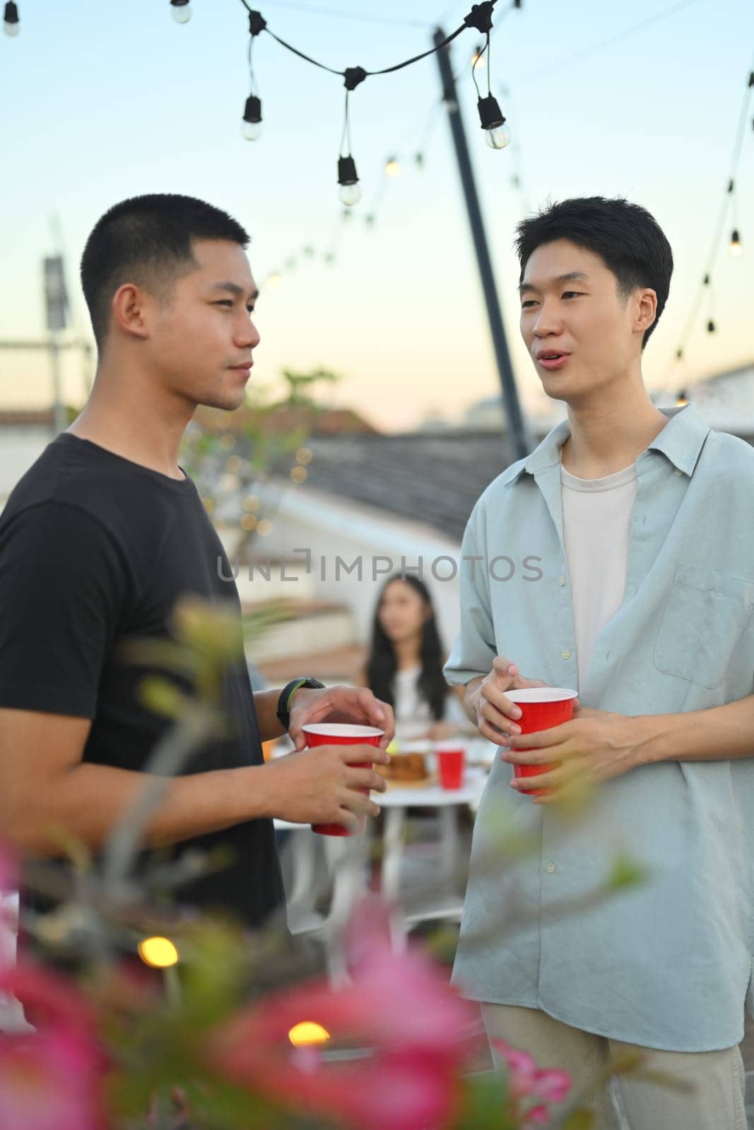 Two young handsome men having drinks and chatting at the rooftop party in the evening. by prathanchorruangsak