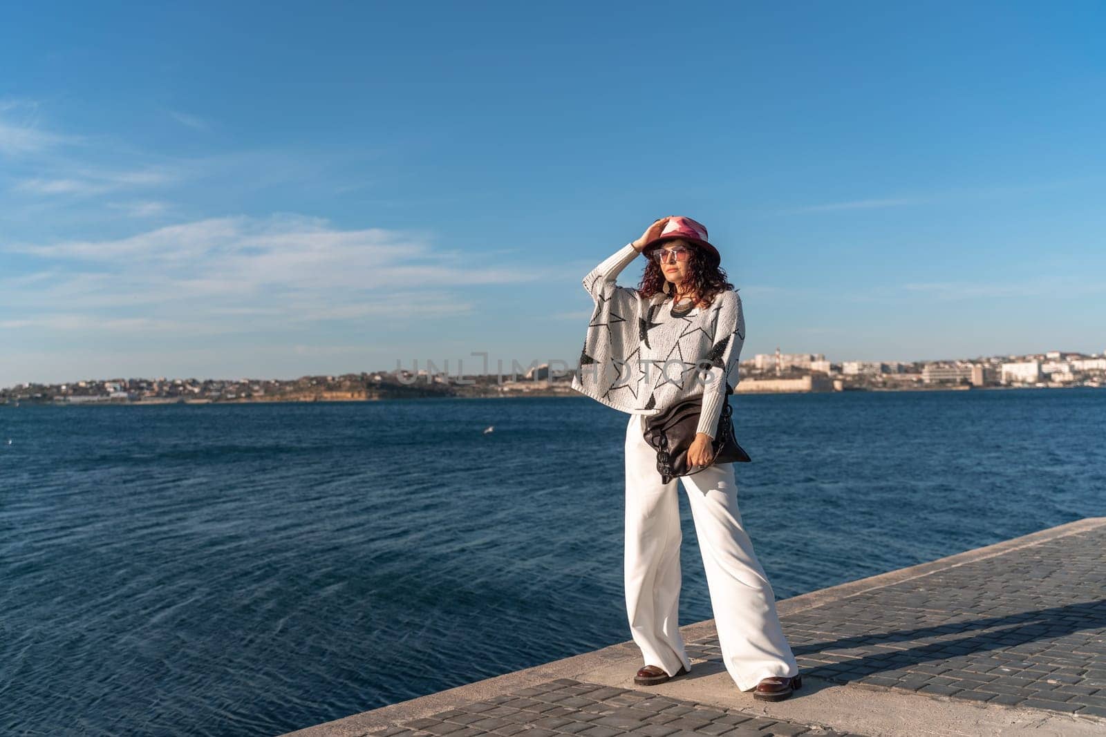 Stylish seashore woman. Fashionable woman in a hat, white trousers and a light sweater with a black pattern on the background of the sea