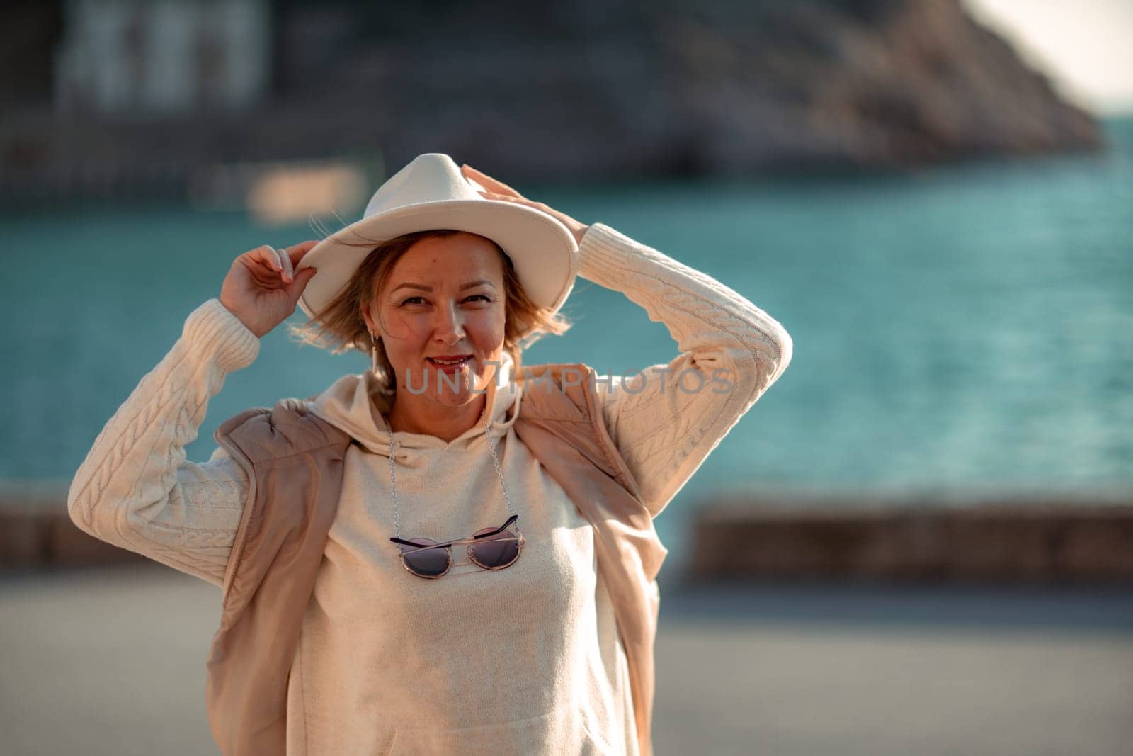 Happy blonde woman in a white suit and hat posing at the camera against the backdrop of the sea.