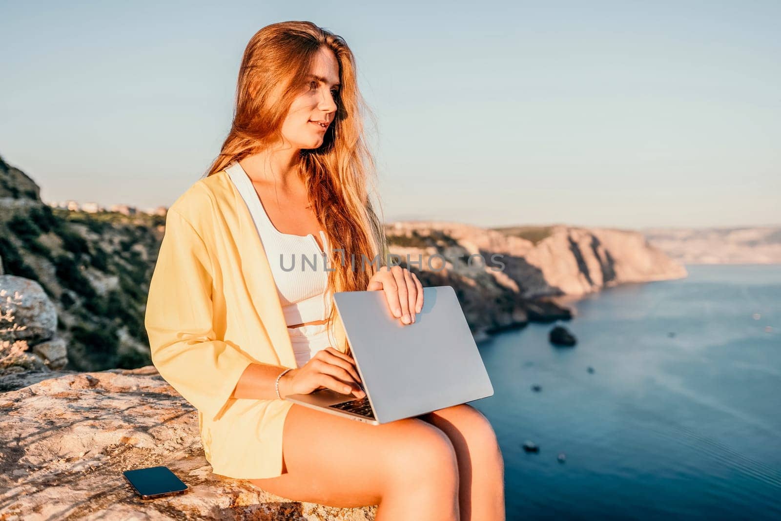 Digital nomad, Business woman working on laptop by the sea. Pretty lady typing on computer by the sea at sunset, makes a business transaction online from a distance. Freelance, remote work on vacation by panophotograph