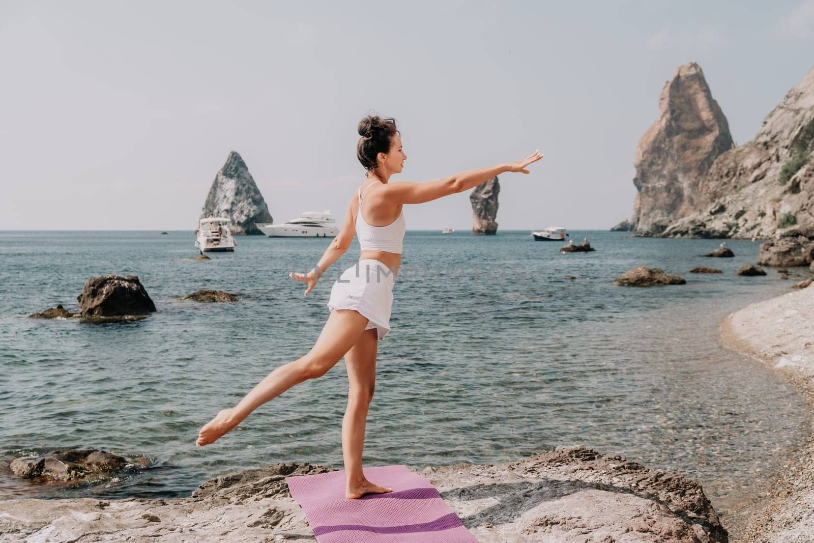 Fitness woman sea. A happy middle aged woman in white sportswear exercises morning outdoors on a beach with volcanic rocks by the sea. Female fitness pilates yoga routine concept. Healthy lifestyle. by panophotograph