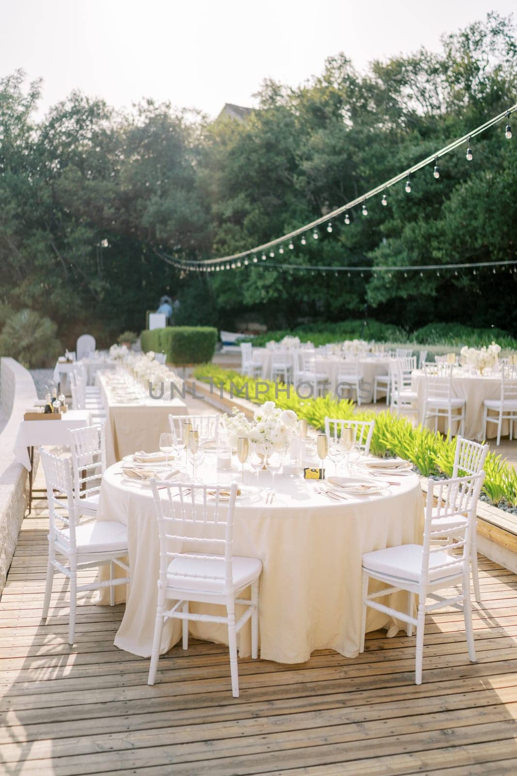 Festive tables with bouquets of flowers stand in the garden on a wooden flooring among the trees. High quality photo