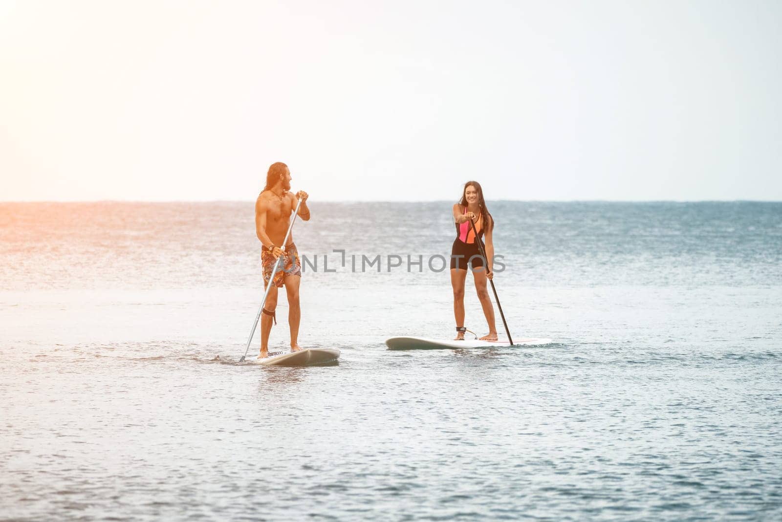 Sea woman and man on sup. Silhouette of happy young woman and man, surfing on SUP board, confident paddling through water surface. Idyllic sunset. Active lifestyle at sea or river