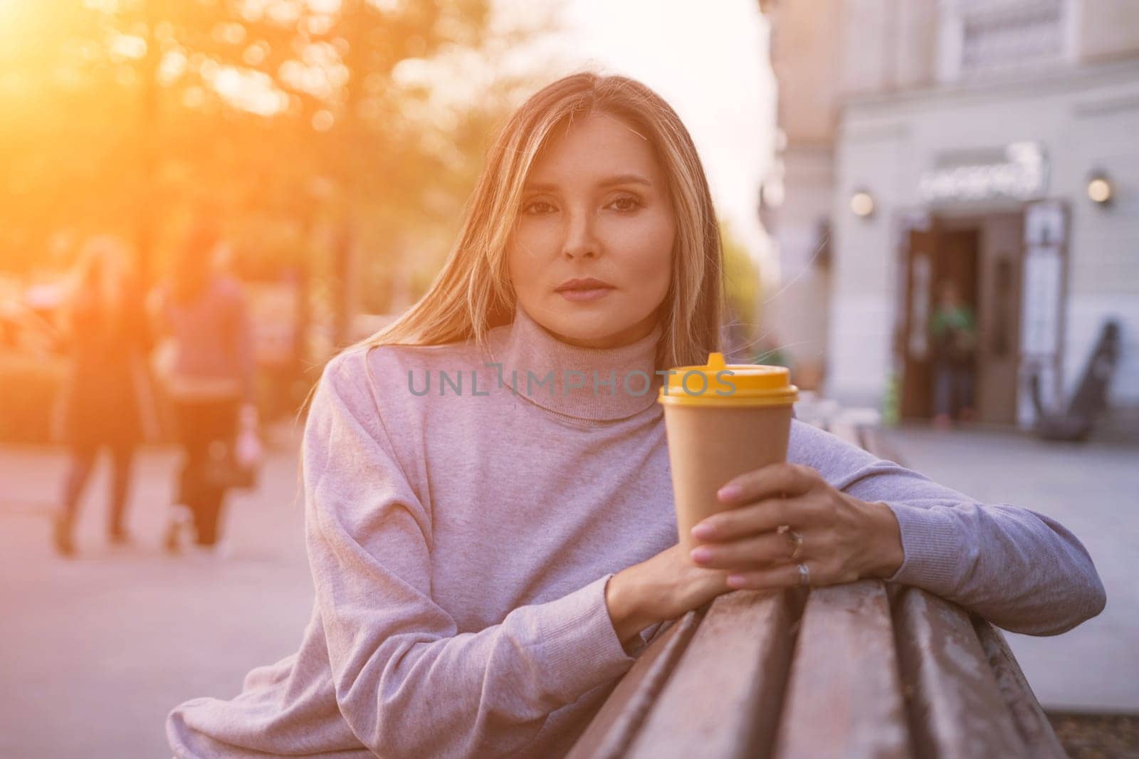 Woman drinks from cup on wooden bench. She is wearing a white shirt enjoying her beverage. The bench is located in a park setting, with trees in the background