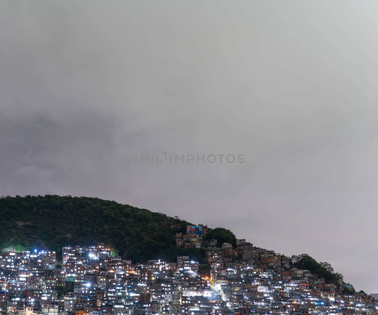 Illuminated favela on a hillside at dusk with compact housing and overcast skies.