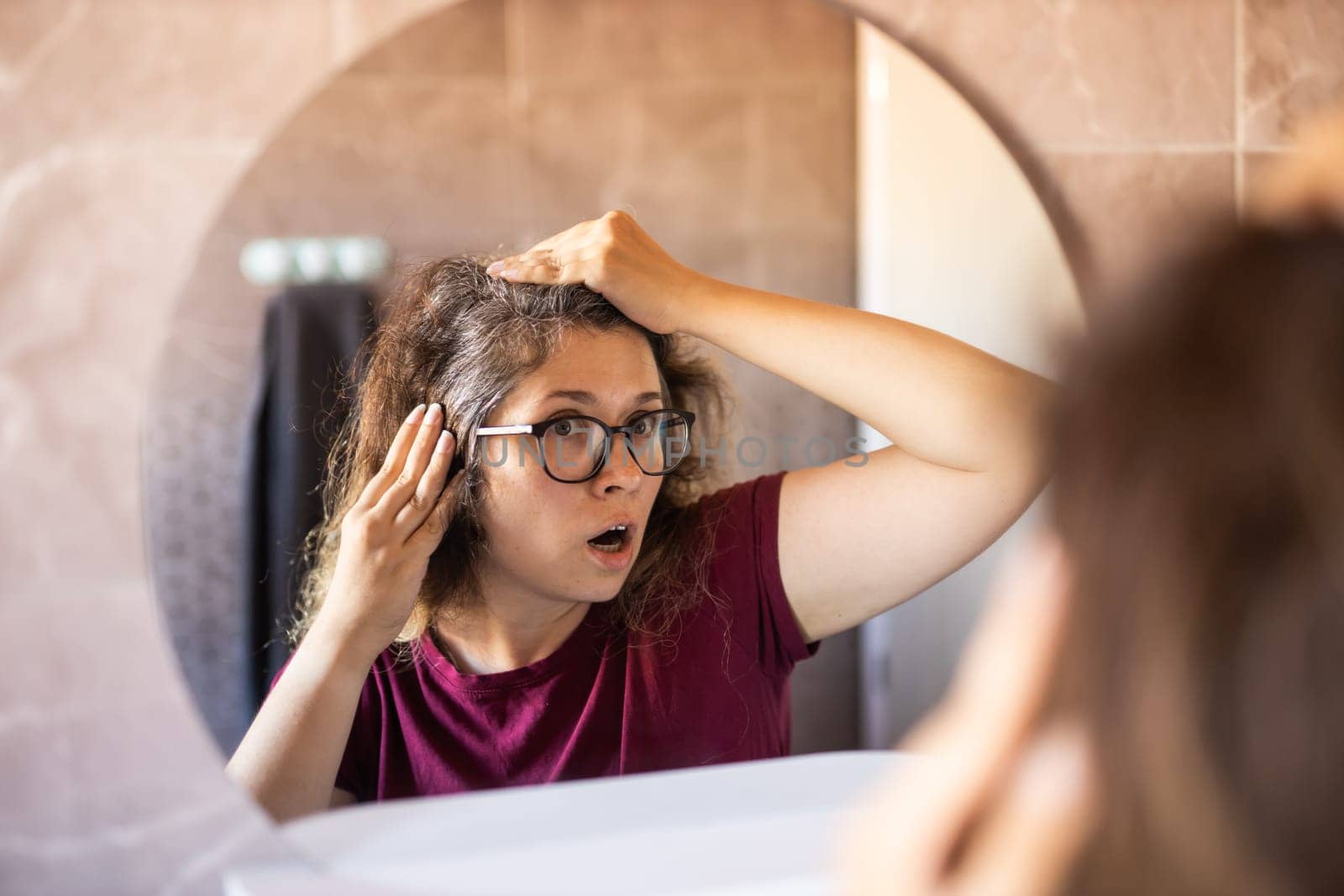 Woman Looking for Gray Hair in the Mirror. Young woman scared of premature aging signs by Satura86