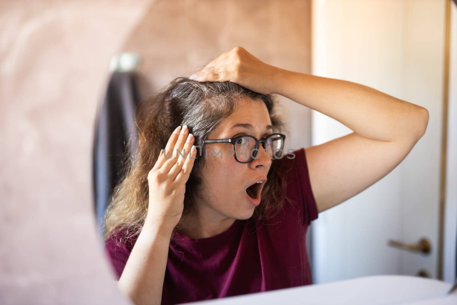 Woman Looking for Gray Hair in the Mirror. Young woman scared of premature aging signs.