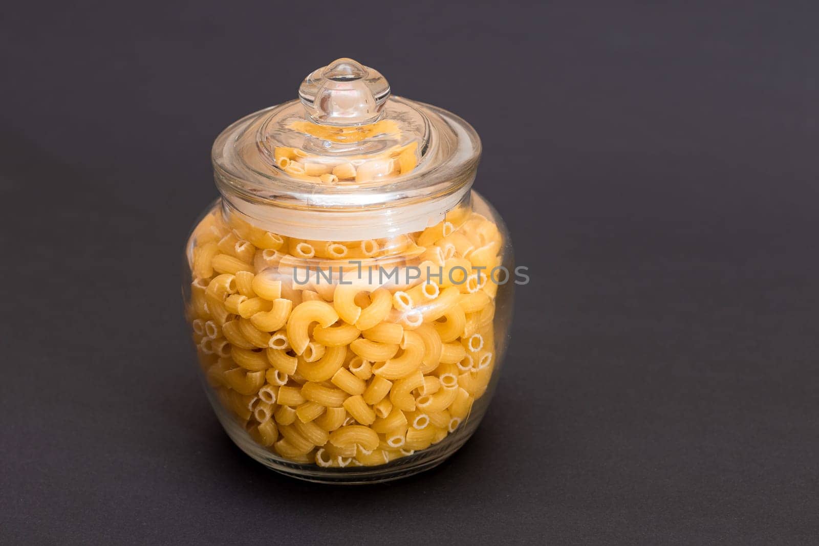 Uncooked Chifferi Rigati Pasta in Glass Jar on Black Background by InfinitumProdux