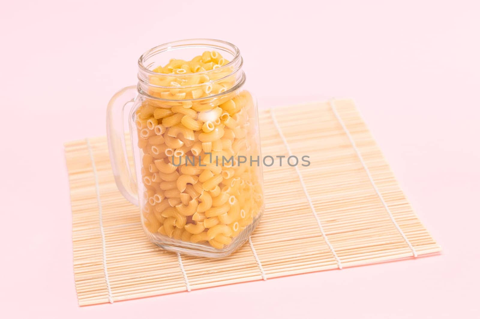 Uncooked Chifferi Rigati Pasta in Glass Jar on Bamboo Mat on Pink Background by InfinitumProdux