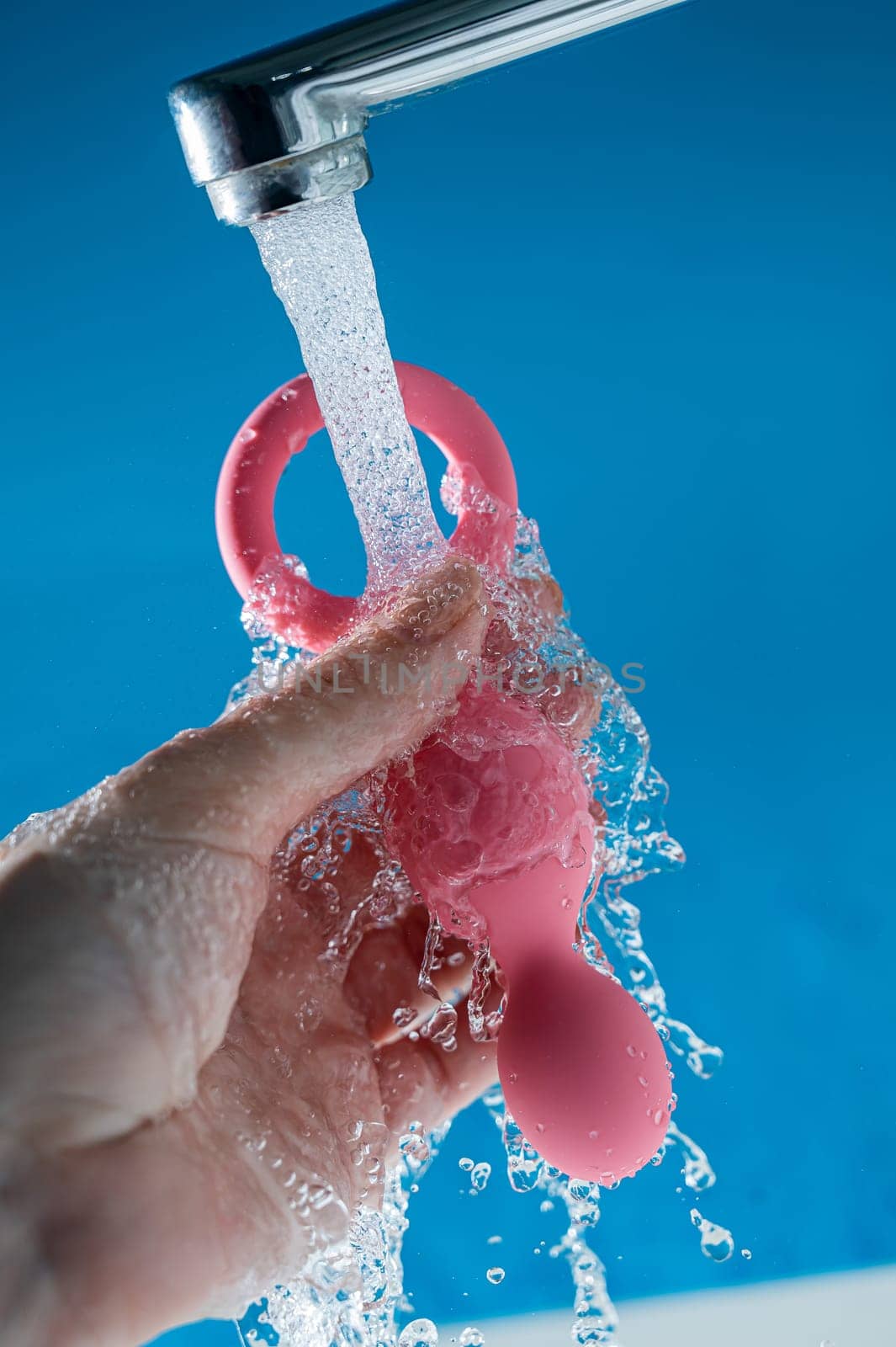 Woman holding pink anal beads under running water on blue background. Sex toy hygiene concept. by mrwed54