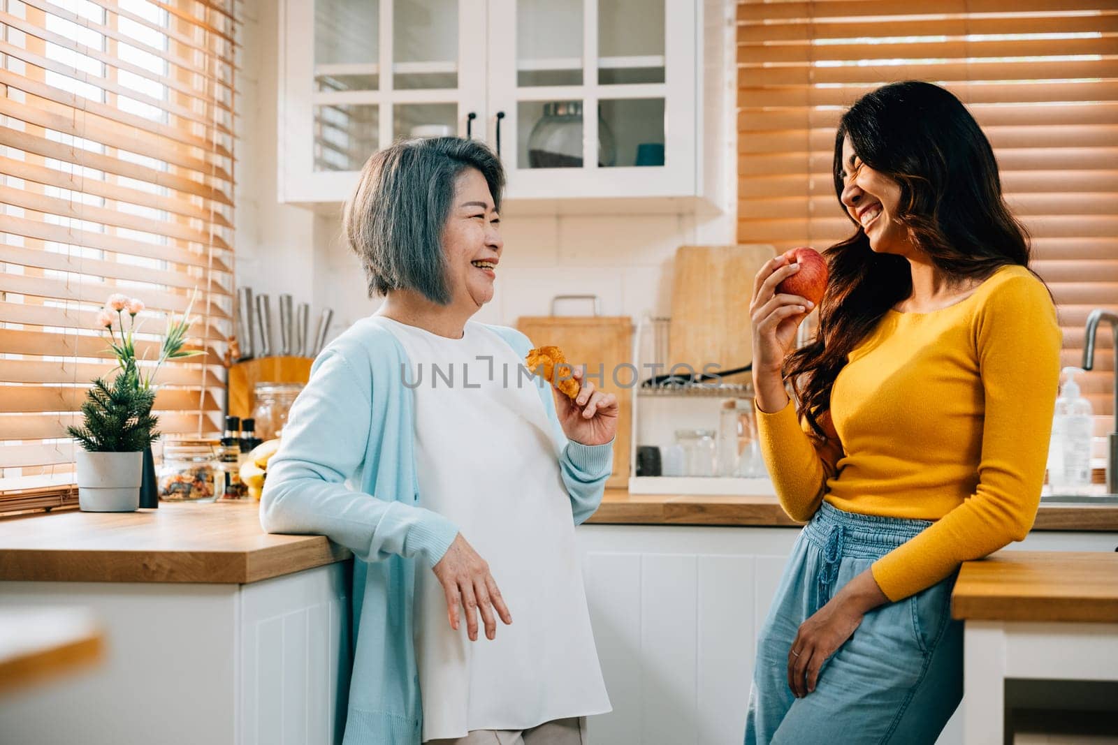 A smiling mother and her adult daughter, an attractive young woman, stand together in the kitchen, holding an apple. Their bond radiates happiness and family togetherness.