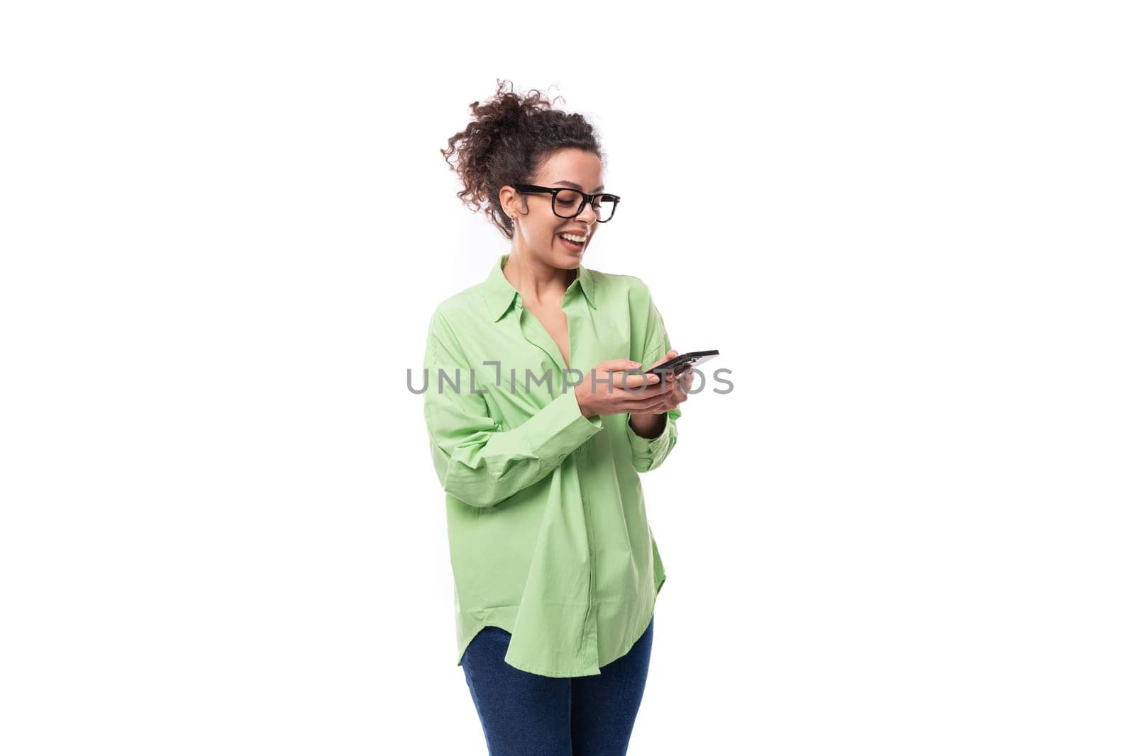 pretty young brunette curly woman in glasses dressed in a green shirt is chatting in a smartphone.