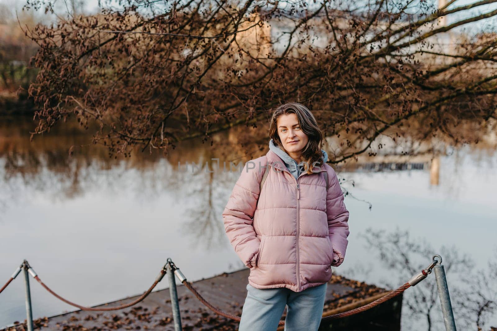 A young woman standing at the shore looking at the river in autumn sunny day. Street view, copy space for text, travel photo. Happy tourist woman on the bank of the river in autumn in warm clothes. Tourists enjoy their vacation, winter season. Romantic look and travel concept. A joyful mood in a caucasian girl. Winter Wonderland: Enchanting Girl by the Riverside in Autumn by Andrii_Ko