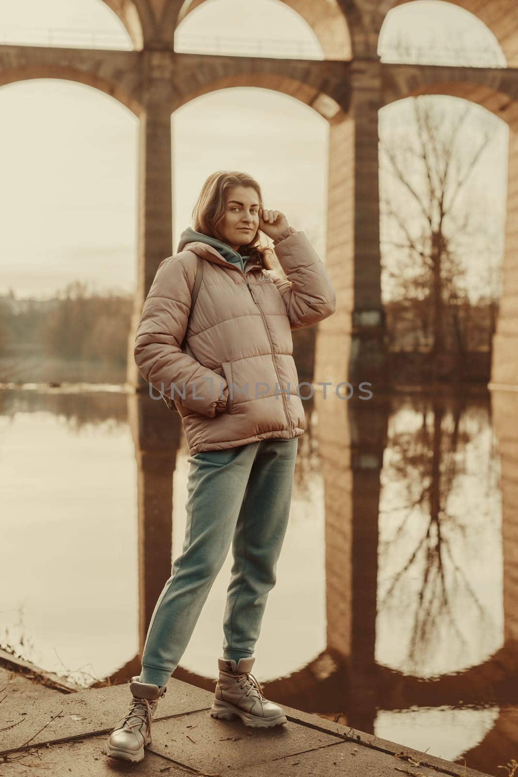 A young woman standing at the shore looking at the river in autumn sunny day. Street view, copy space for text, travel photo. Happy tourist woman on the bank of the river in autumn in warm clothes. Tourists enjoy their vacation, winter season. Romantic look and travel concept. A joyful mood in a caucasian girl. Winter Wonderland: Enchanting Girl by the Riverside in Autumn by Andrii_Ko