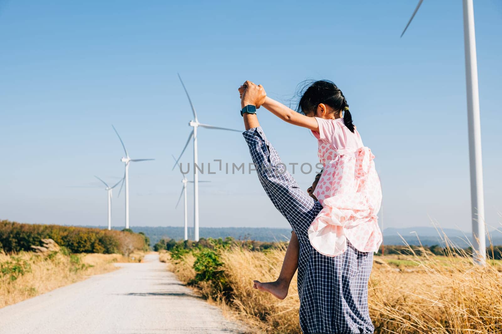 Father joyfully carries daughter exploring wind farm by Sorapop