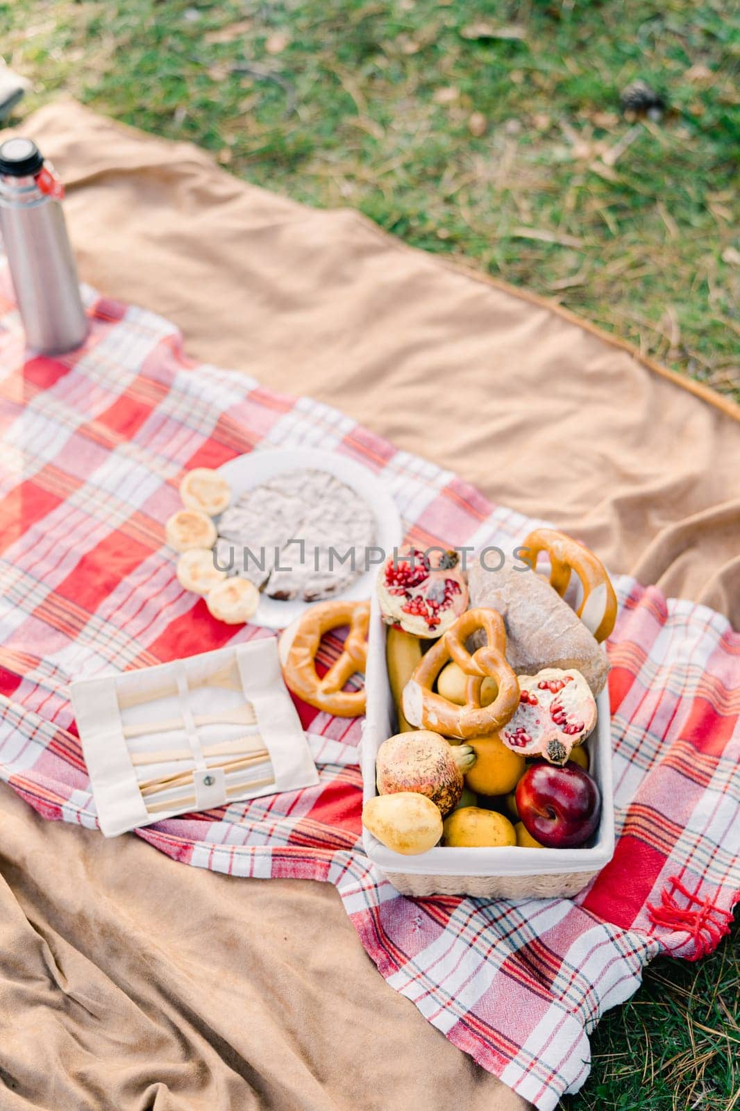 Basket of buns and fruit stands on a blanket next to cutlery and a thermos by Nadtochiy