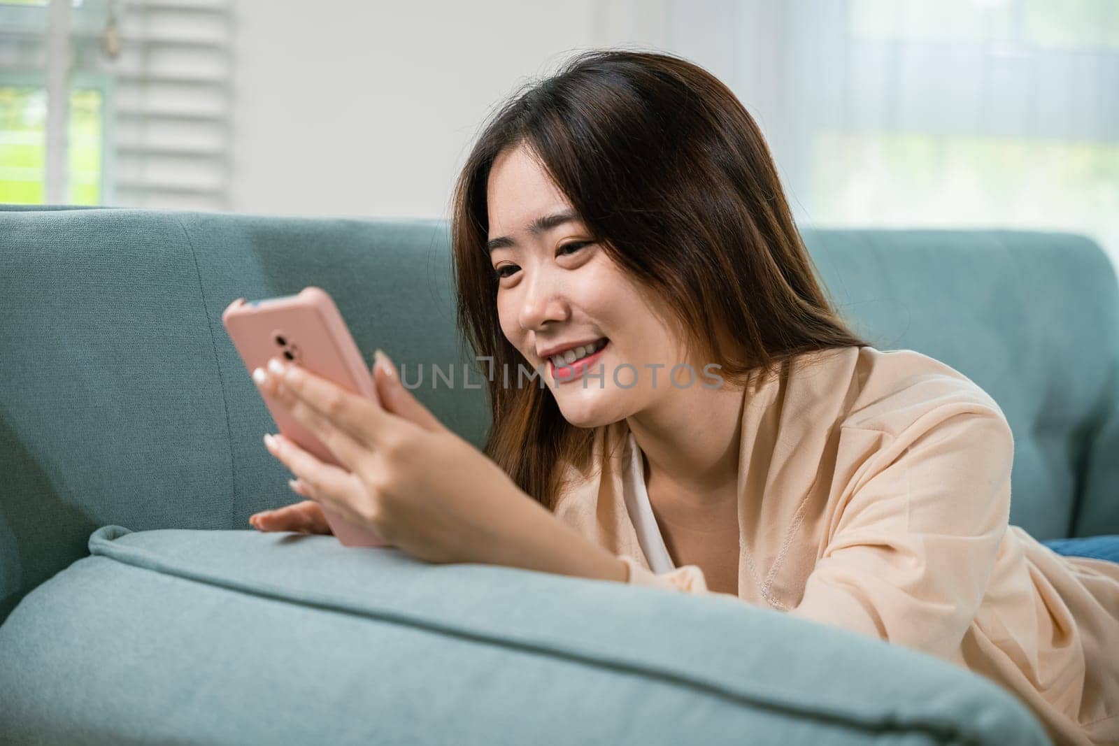 Smiling attractive woman relaxed reading text message on smartphone, Asian young female using smart mobile phone lying on sofa in living room room at home, gadget online application making internet
