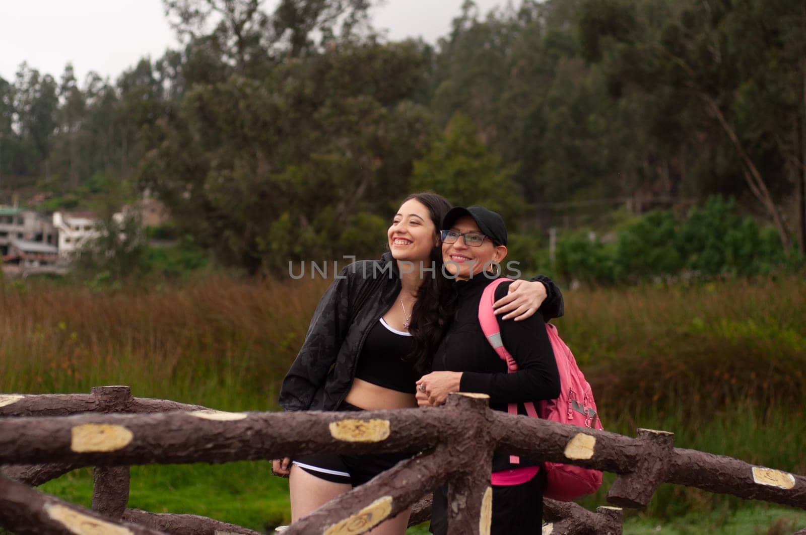 mother and daughter dressed in sportswear and a pink backpack embraced looking at the horizon on a wooden bridge. women's day by Raulmartin