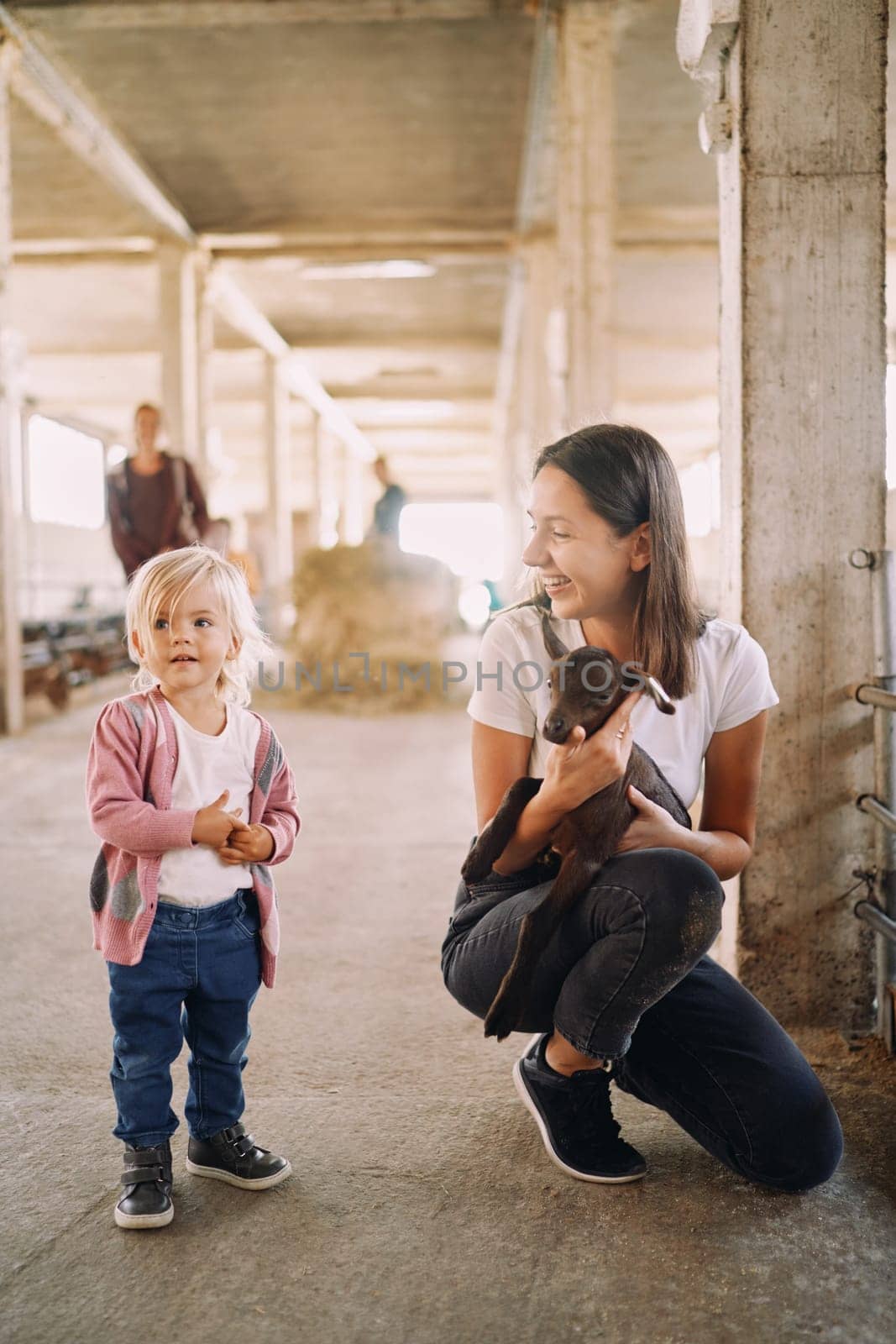 Squatting mother with a goatling in her arms looks at a little girl standing nearby. High quality photo