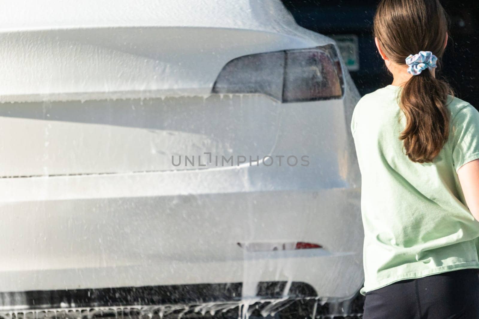 A young girl enthusiastically assists in washing the family's electric car in their suburban driveway.