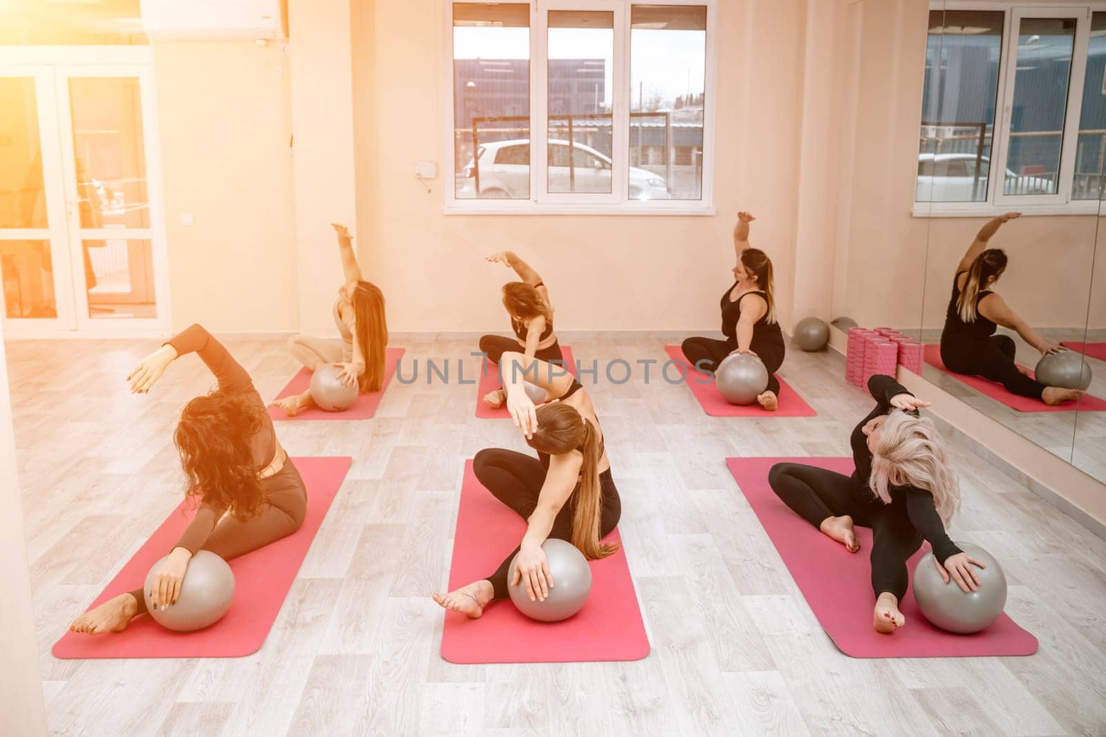 A group of six athletic women doing pilates or yoga on pink mats in front of a window in a beige loft studio interior. Teamwork, good mood and healthy lifestyle concept. by Matiunina