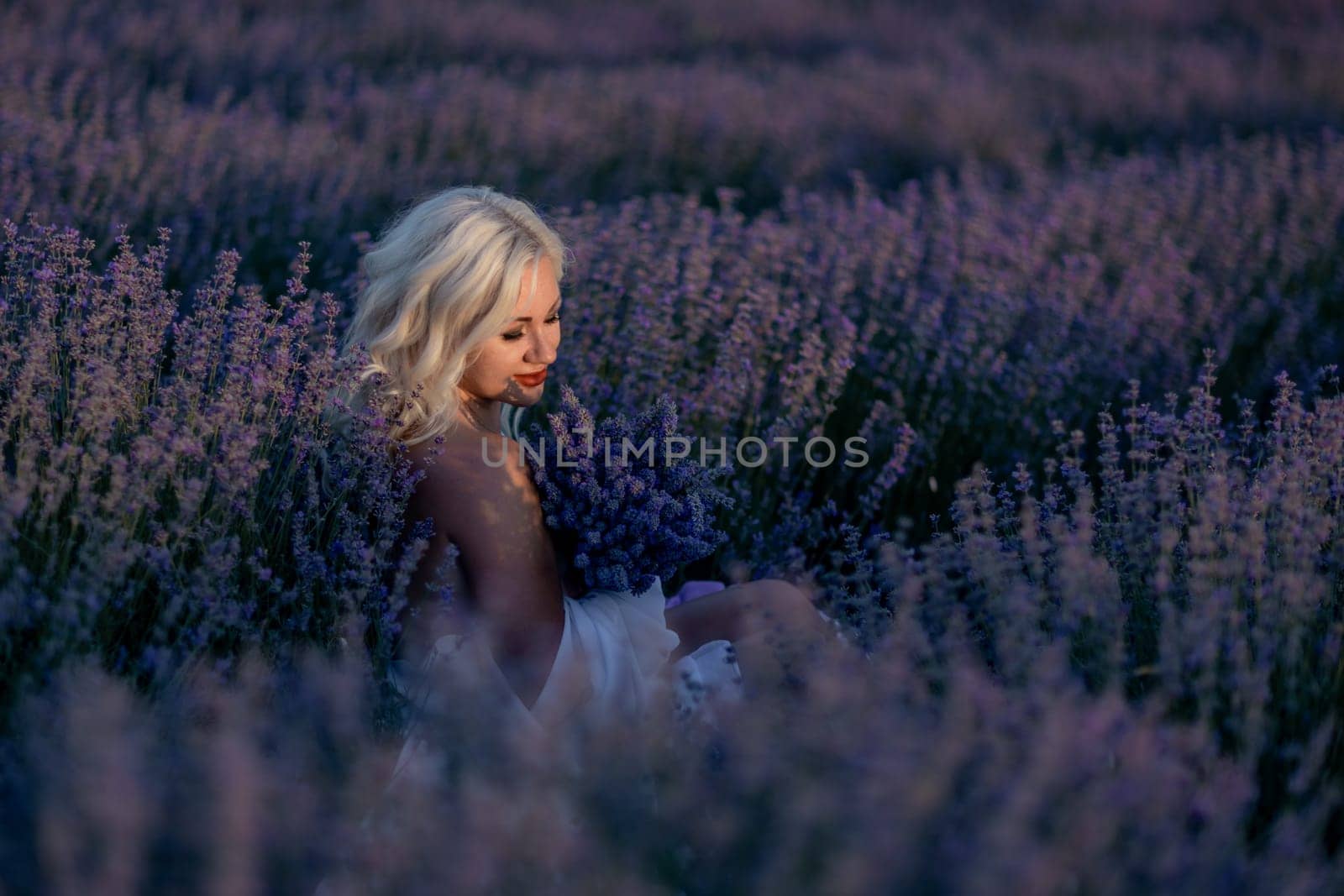 Blonde woman poses in lavender field at sunset. Happy woman in white dress holds lavender bouquet. Aromatherapy concept, lavender oil, photo session in lavender.
