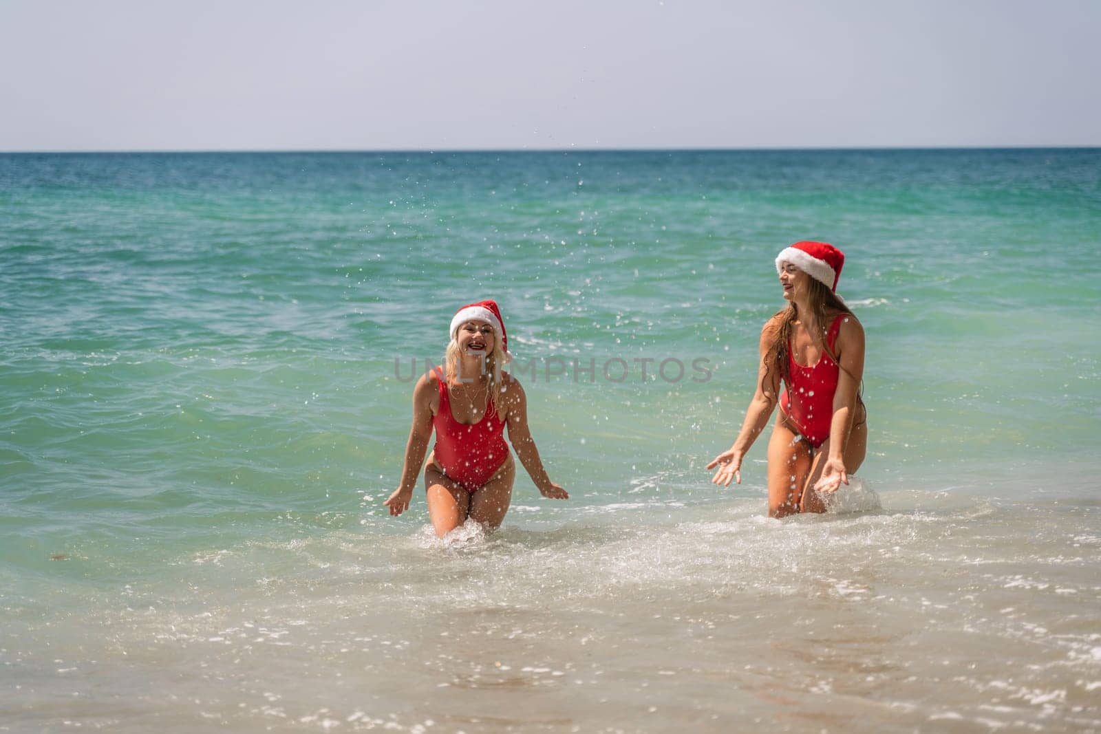 Women Santa hats ocean play. Seaside, beach daytime, enjoying beach fun. Two women in red swimsuits and Santa hats are enjoying themselves in the ocean waves