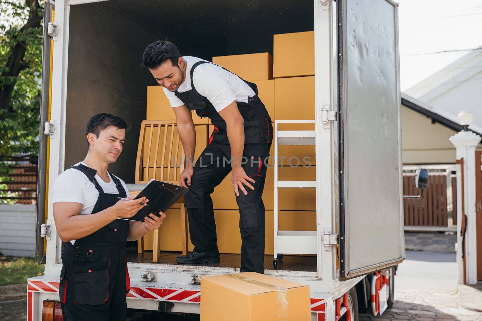 Mover workers in uniform unload boxes checking checklist with a clipboard at the truck. Professional delivery service ensures smooth relocation and efficient shipping. Moving day concept